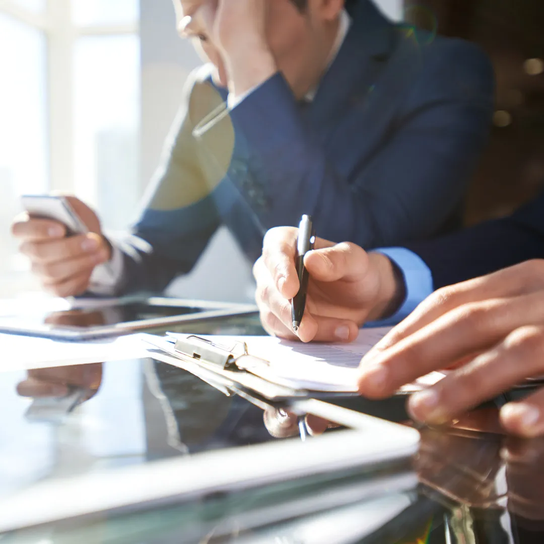 Bilde av et bord i konferanserom der du ser flere hender med pen og pc, uklare ansikter 